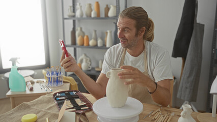 Man holding smartphone while pointing finger to ceramic vase in pottery studio; creativity focus...