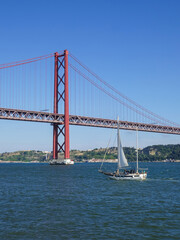 Obraz premium Graceful sailboat with tall white sails moving through vivid blue waters under a cloudless sky, framed by the striking lines of a red suspension bridge.