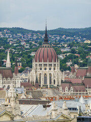Fototapeta premium Majestic parliament building with a prominent dome and decorative façade, standing out over rooftops and framed by a green hillside beneath overcast light.