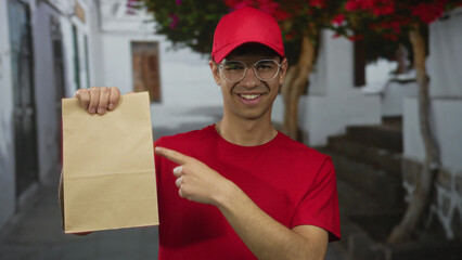 Young hispanic delivery man in red shirt and cap holds a paper bag and points finger to bag on...