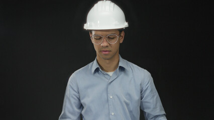 Man engineer wearing white hardhat and clear glasses reading large blueprint sheet in studio; concentration.
