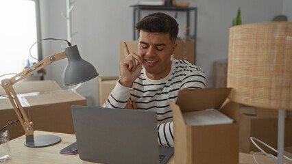 Young man in a striped shirt sitting at a desk in a new home office surrounded by boxes, looking...