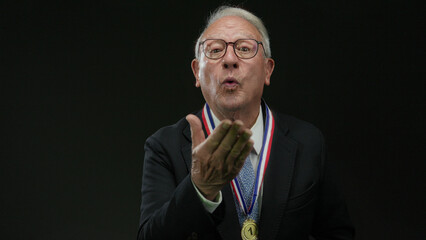 Senior man in a suit with a medal blows a kiss against a black background, conveying celebration...