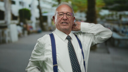 Senior man in business suit stands outdoors on a street gesturing confidently while wearing glasses...