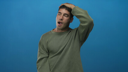 Young hispanic man wearing olive green sweater holds hand to forehead and opens mouth in studio;...
