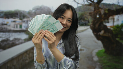 Young woman smiling outdoors holding chinese yuan banknotes on a seaside promenade with a backdrop...