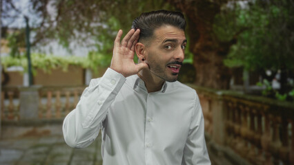 Man in white shirt cups his ear while standing by a stone balustrade along a pathway in green park;...