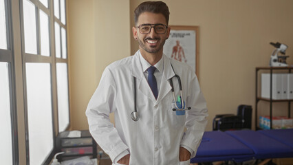 Man with stethoscope crossing arms in bright clinic wearing white coat and tie; professional...