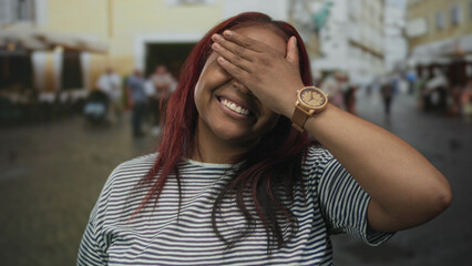 Woman covers her eyes with a raised hand while smiling warmly under bright daylight on a bustling...