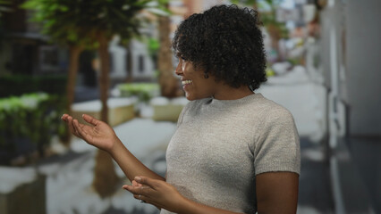 Smiling young african american woman with open palm by parked cars on busy street; friendly...