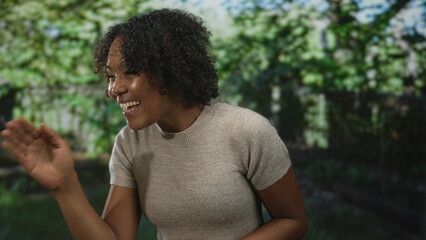 Young african american woman in sunlit green park smiling and laughing with raised hands wearing...