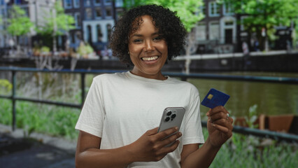 Woman holds smartphone and creditcard by a canal railing while smiling broadly on a city street; confidence.