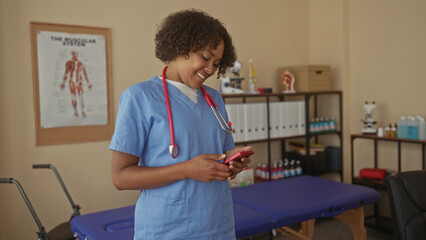 Obraz premium Young african american woman doctor scrolls smartphone in exam room beside anatomical chart and treatment table; concentration.