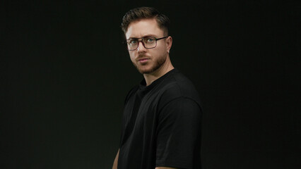 Obraz premium Young man with beard and glasses stands confidently against a black wall with an isolated and focused demeanor.