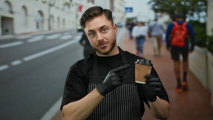 Young man with beard in apron points at coffee cup while standing on city street, highlighting his...