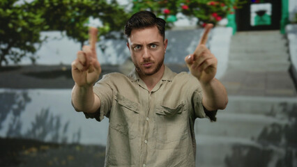 Young man with beard signaling no with fingers on city street lined with greenery portraying urban...