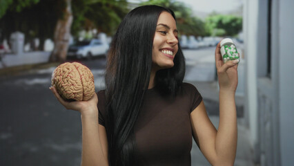 Woman smiling happily on a city street holding a model brain and bottle of pills, showcasing a juxtaposition of natural intelligence and artificial enhancement.