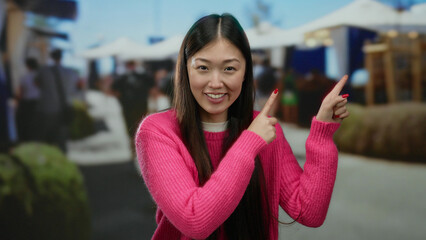 Woman wearing pink sweater pointing outdoors at a bustling market scene, surrounded by people and...