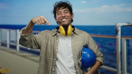 Fototapeta premium Hispanic man smiling on a cruise ship deck, wearing noise-canceling headphones and holding a blue hardhat with the ocean in the background.