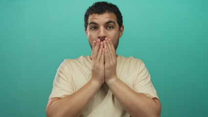 Man holds cheeks with both hands and opens mouth wide against a turquoise studio wall for a...