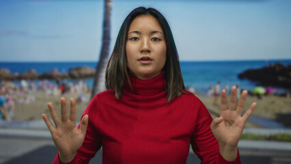 Woman gesturing calm at the beach in a red sweater with a blurred seaside background, conveying...
