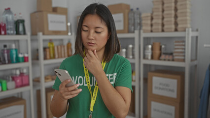 Woman in green volunteer shirt uses smartphone in donation center surrounded by organized supplies...