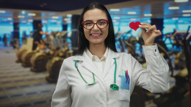 Young woman doctor with stethoscope holding heart in gym setting, showcasing health awareness among medical professionals in athletic environments with modern sports equipment.