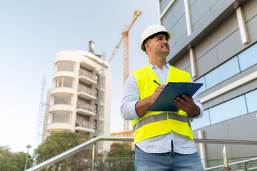 Construction Engineer in hardhat writing notes on clipboard at construction site inspecting building