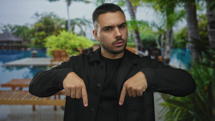 Young hispanic man pointing both index fingers down at poolside building walkway outdoors;...