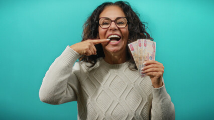 Middle-aged woman in glasses holding uae dirhams against blue wall, smiling and pointing at her...