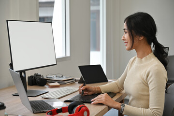 Asian woman using graphics tablet to design work on computer with blank screen on desk full of...