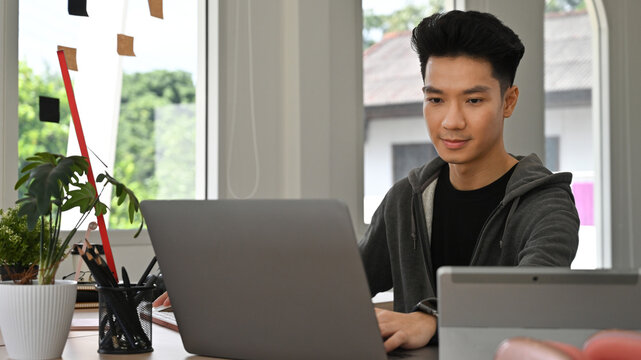 A Focused Asian Creative freelancer man works on a laptop, developing graphic content from an office workspace - Powered by Adobe