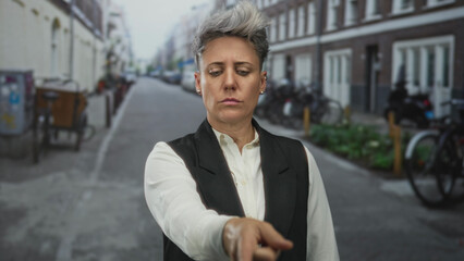 Caucasian woman with short hair and neutral expression pointing finger to street lined with brick...