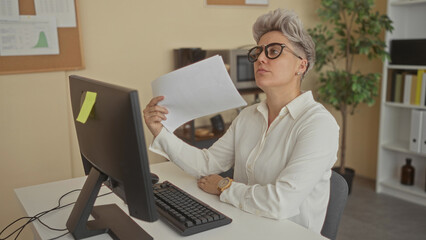 Woman wearing glasses fans face with documents inside modern office building by computer desk;...