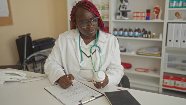 Woman doctor in white coat writing notes and holding medicine bottle in clinic room with shelves in background suggesting medical professional setting.