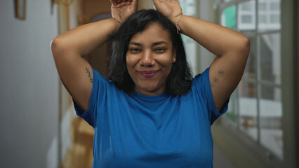 African american woman in blue shirt holds hands on head and smiles in hotel building corridor;...