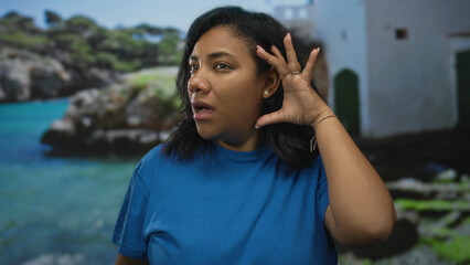 Woman with neutral expression cups ear while standing in studio against seaside backdrop and wearing blue shirt with direct gaze at camera; curiosity.