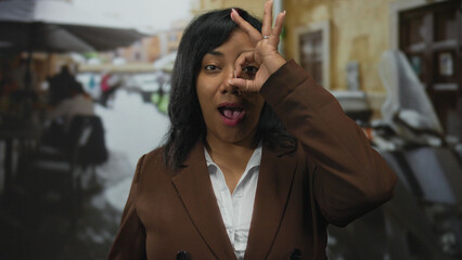 African american woman in brown blazer and white shirt makes ok sign to eye on street; playfulness...