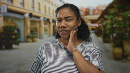 African american woman touches cheek with hand in sunlit busy street lined with shops and potted...