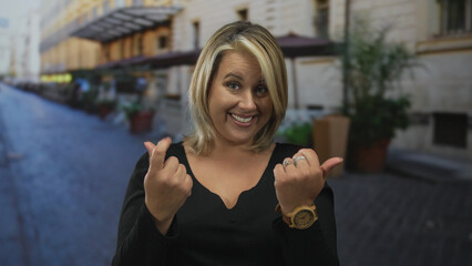 Young blonde woman pointing fingers forward in a city street with blurred buildings behind her;...