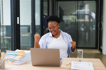 Fototapeta premium Young african american businesswoman is raising her fists in excitement while sitting at her desk in a modern office, celebrating a successful business deal or project completion