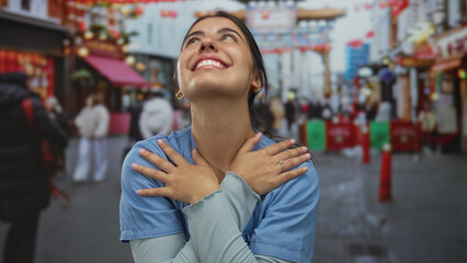 Woman in blue medical scrubs hugging herself on a street with blurred pedestrians and red lanterns...