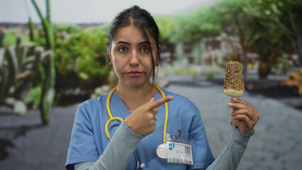 Young hispanic woman doctor in blue uniform points finger at ice cream bar in green park; healthy...
