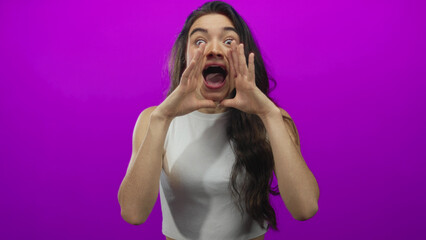 Hispanic young woman cups hands around wide open mouth shouting in purple studio; announcement...