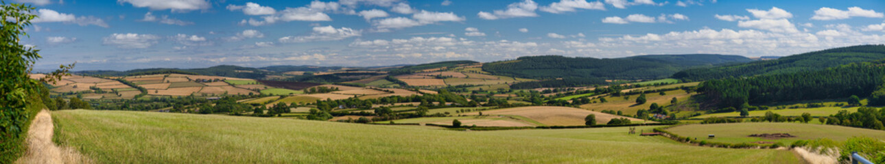 Wide panoramic view of patchwork fields and rolling hills under blue skies in the Shropshire Hills National Landscape, Shropshire, UK. Bright summer light and fluffy clouds convey a fresh rural feel.