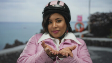 Young woman wearing helmet and pink jacket extends hands towards camera by the seaside promenade