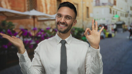 Hispanic man smiling in an urban street setting, making an okay gesture and extending his hand,...