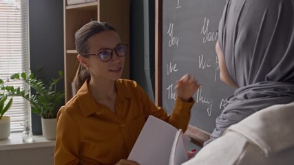 In multicultural classroom, Caucasian female teacher guiding Muslim student through English pronouns using written examples on chalkboard - Powered by Adobe