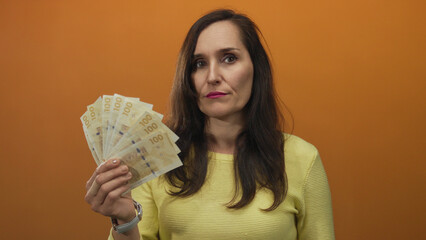 Woman showing swedish kronor with serious expression against orange wall background, emphasizing...