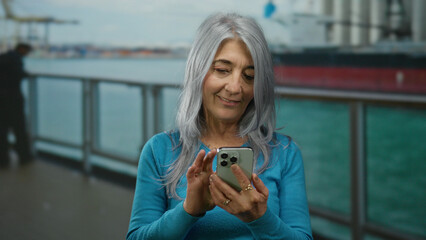Senior woman with grey hair using phone on a cruise boat against seaside backdrop, enjoying a tranquil moment outdoors with a candid smile.